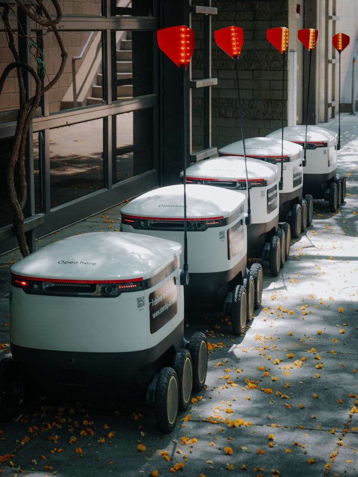 gallery-1 Line of autonomous food delivery robots on urban path under trees.
